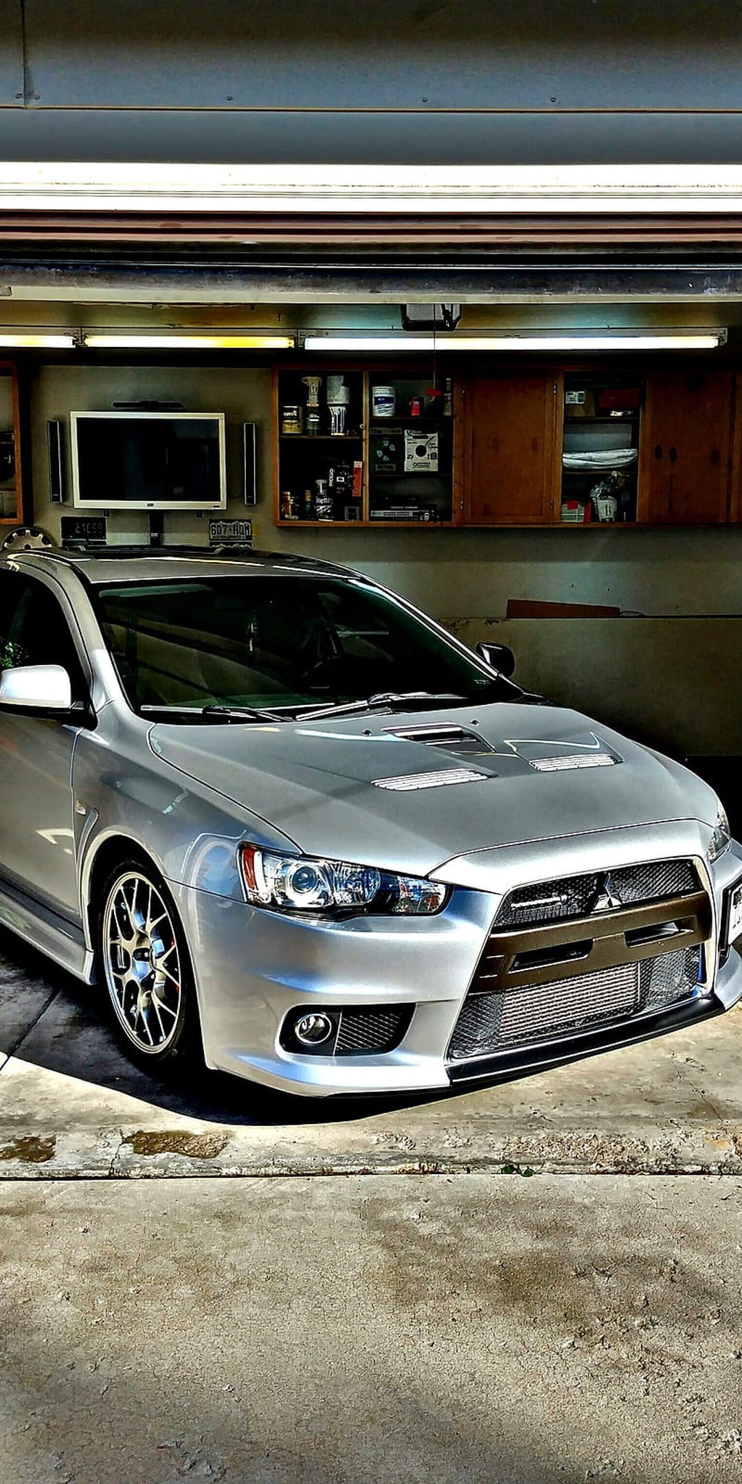 Silver Mitsubishi Lancer Evo X parked in a bright garage featuring hood vents and dark aftermarket wheels.