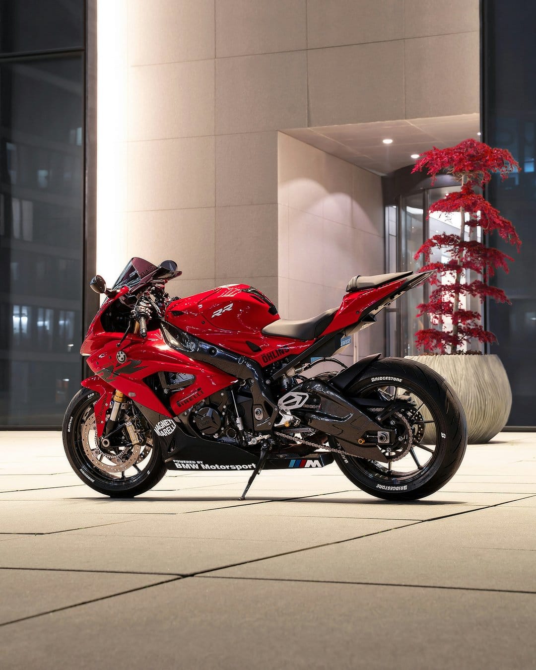 Red BMW S 1000 RR motorcycle parked on tiled floor next to a potted maple tree and modern building at night.