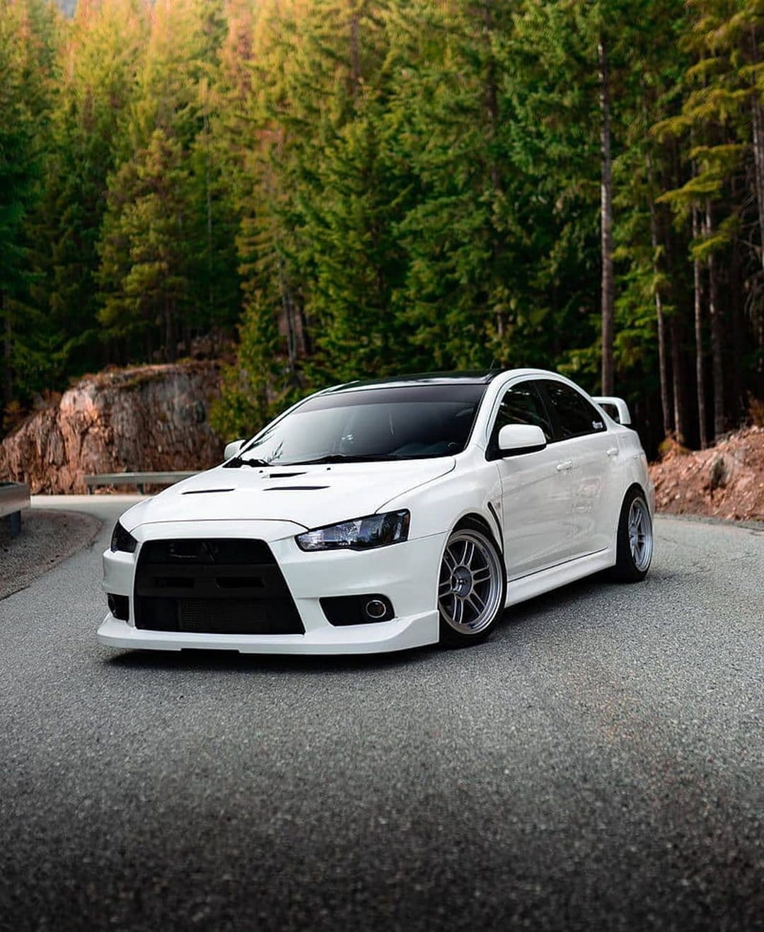 White Mitsubishi Lancer Evolution driving on a winding asphalt road through a dense forest of evergreen trees.