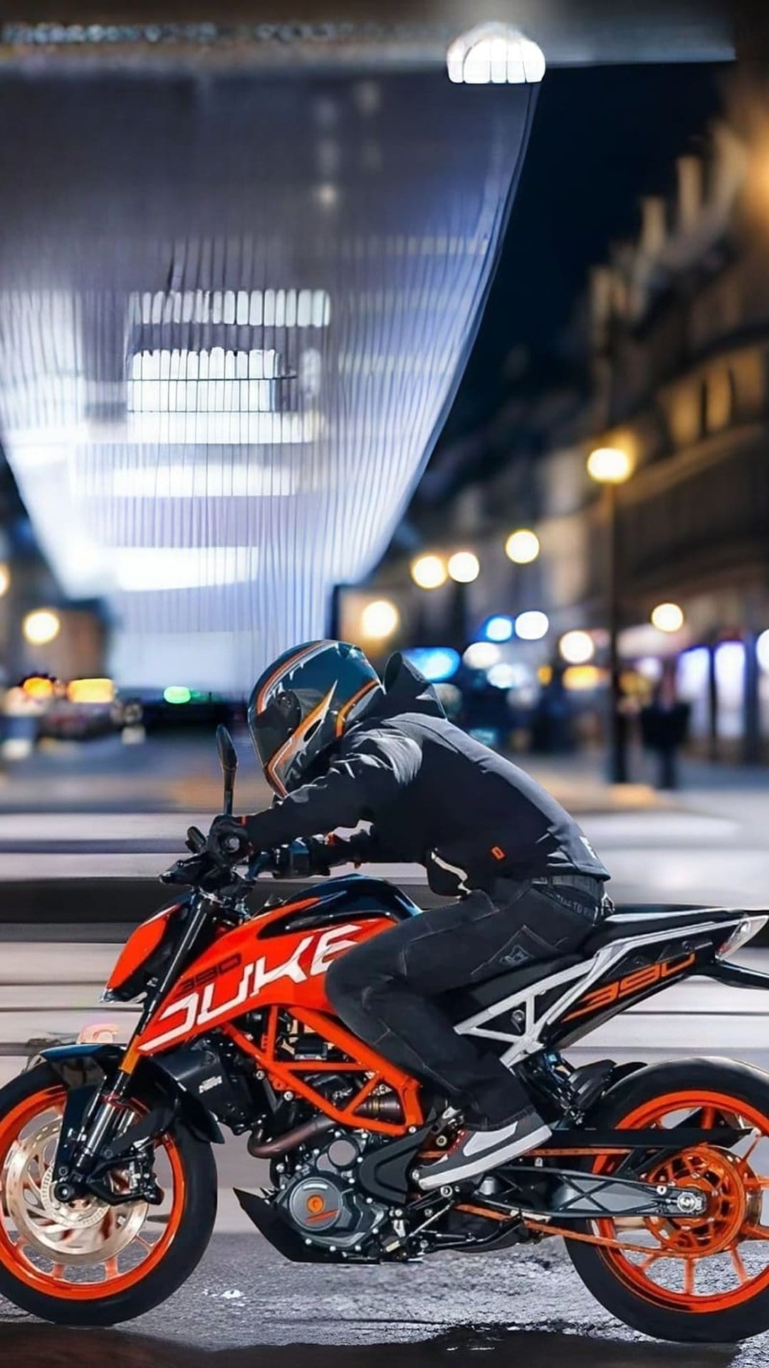 Motorcyclist on an orange KTM Duke 390 cruises under glowing urban structures amidst blurred city night lights.