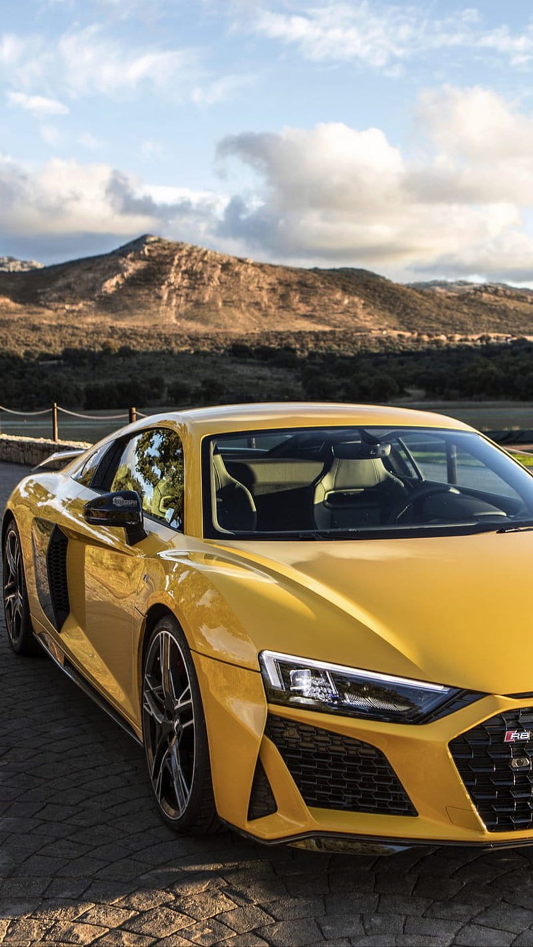 Yellow Audi R8 parked on a textured cobblestone driveway with rolling hills and a cloudy sky in the background.