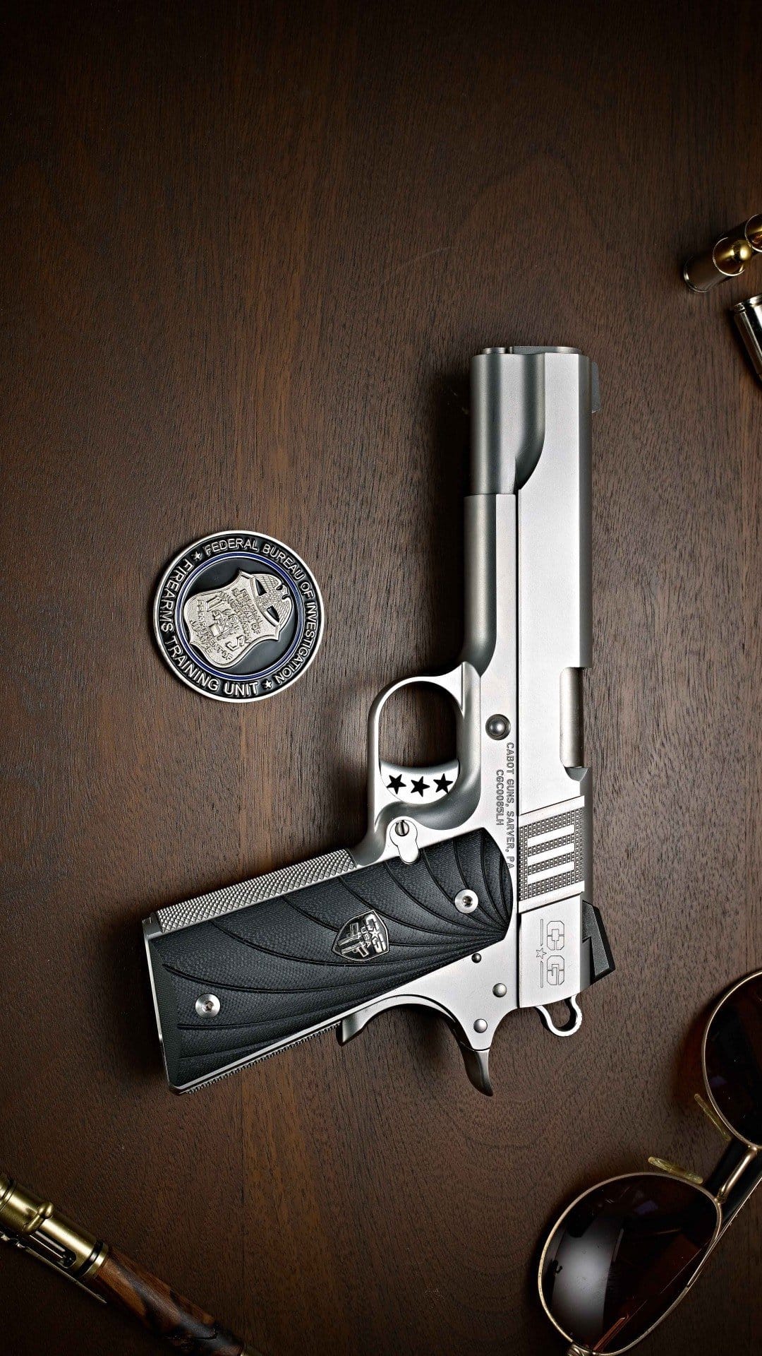 Silver semi-automatic pistol, law enforcement coin, and sunglasses rest on a dark textured wooden background.