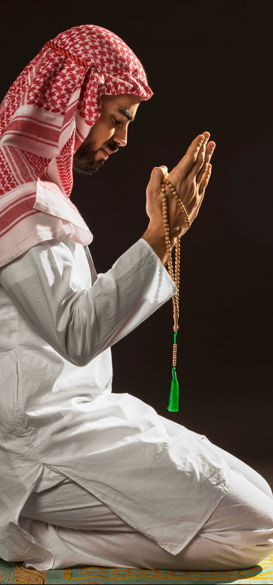 Man in a white thobe and red keffiyeh holds wooden prayer beads while kneeling in a dark, atmospheric setting.