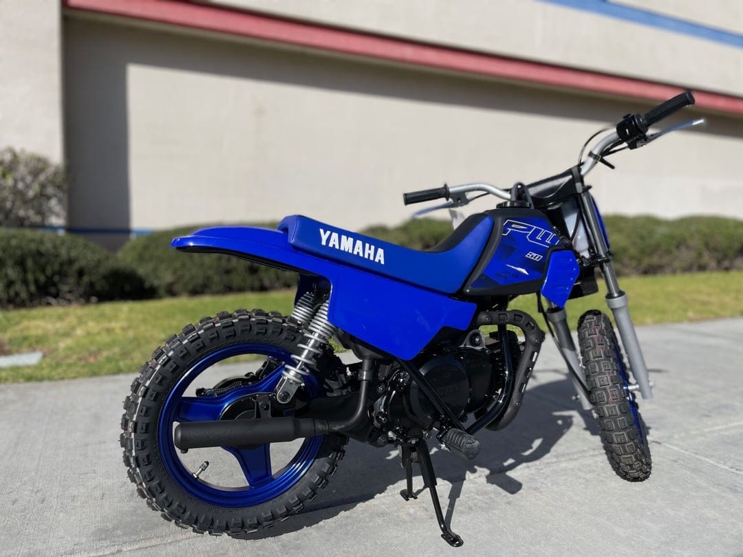 Blue Yamaha PW50 dirt bike parked on concrete with a tan building and green bushes in the background.