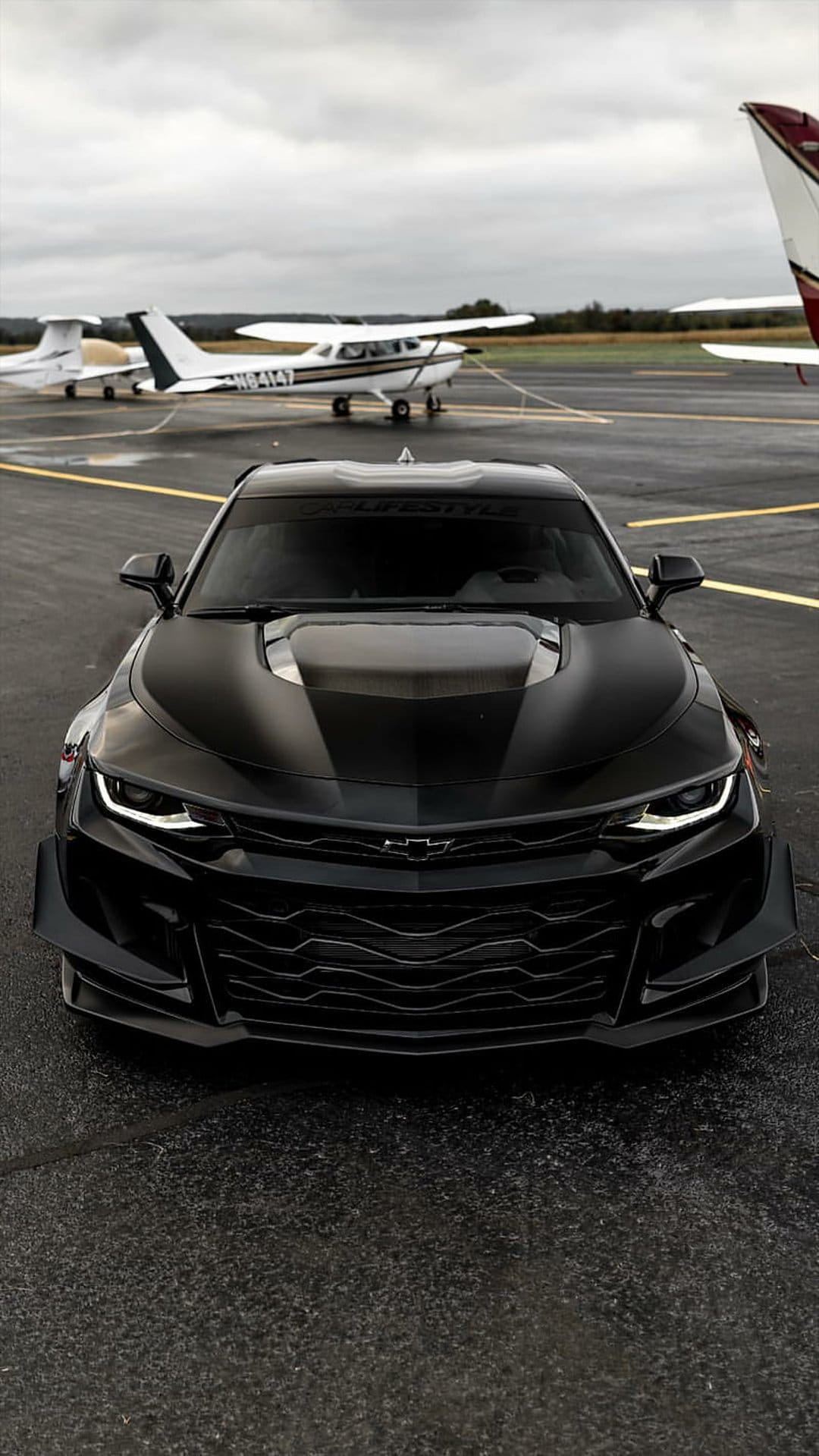 Black Chevrolet Camaro parked on a damp airport runway near propeller planes under a heavy overcast sky.