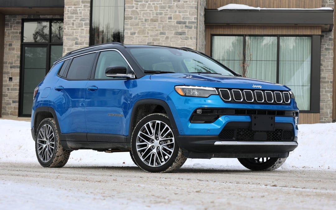 Blue Jeep Compass SUV parked on a snowy road in front of a modern stone building during a bright winter day.
