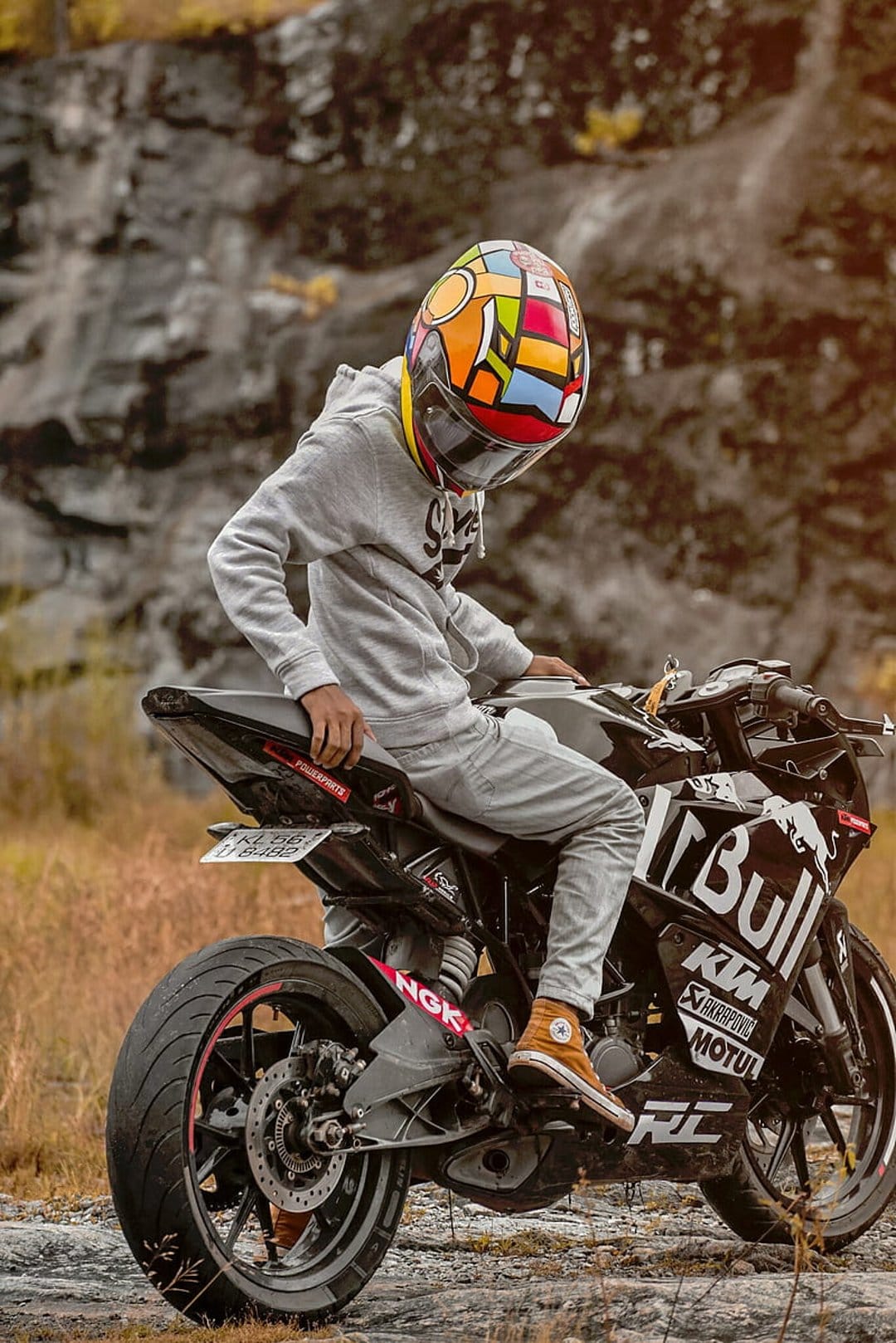 Motorcyclist in a colorful helmet sits on a black KTM bike against a rocky off-road background with dry grass.