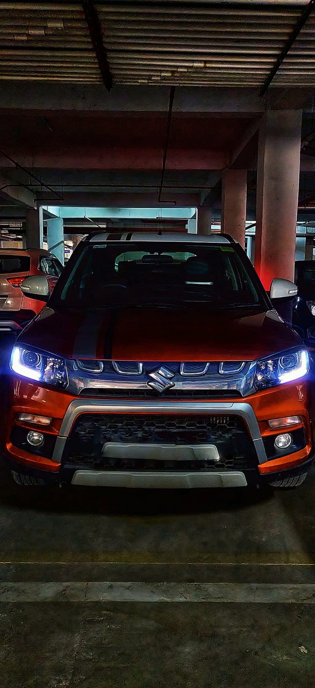 Orange Vitara Brezza SUV with lit headlights and hood stripes parked in a dim concrete underground garage.