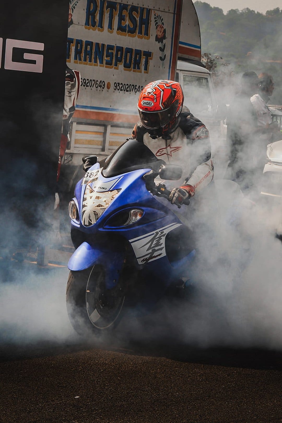 Suzuki Hayabusa motorcycle rider in a white racing suit performs a smoky burnout next to a transport truck.