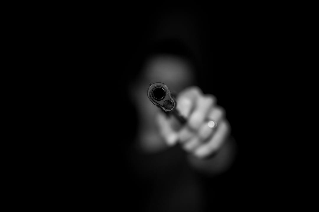 Handgun barrel in sharp focus points at the viewer with a blurred figure in a dark monochrome setting.