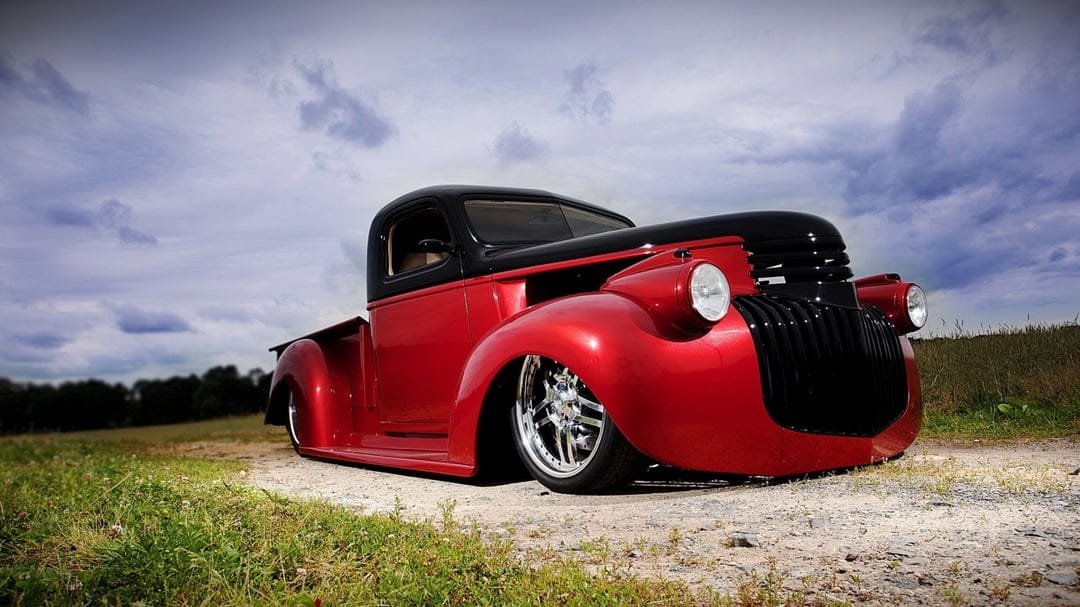 Slammed red and black custom pickup truck with chrome rims on a gravel path under a dramatic, cloudy sky.