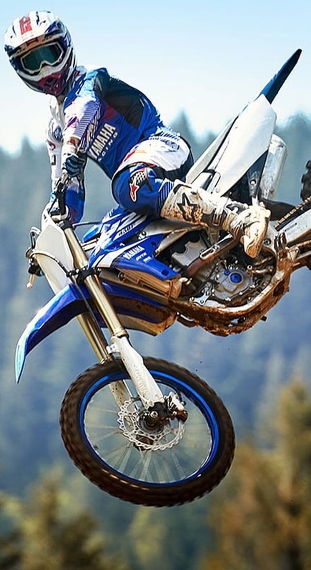 Motocross rider on a blue Yamaha dirt bike jumps mid-air against a blurred green forest background in full gear.