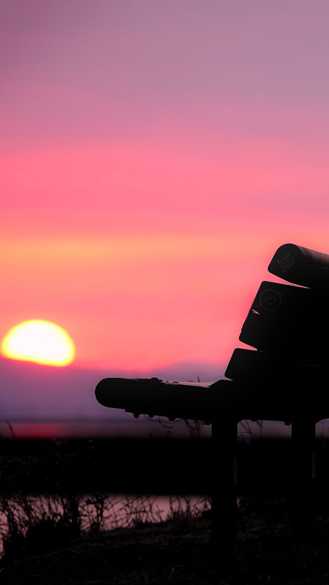 Park bench silhouette sits before a vivid pink and orange sunset sky reflecting on a quiet lake surface.