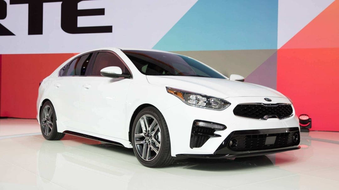 White Kia Forte sedan parked on a reflective showroom floor in front of a colorful geometric background wall.