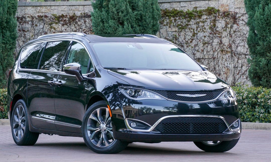 Black Chrysler Pacifica minivan parked on a paved driveway in front of a stone wall and green trees.