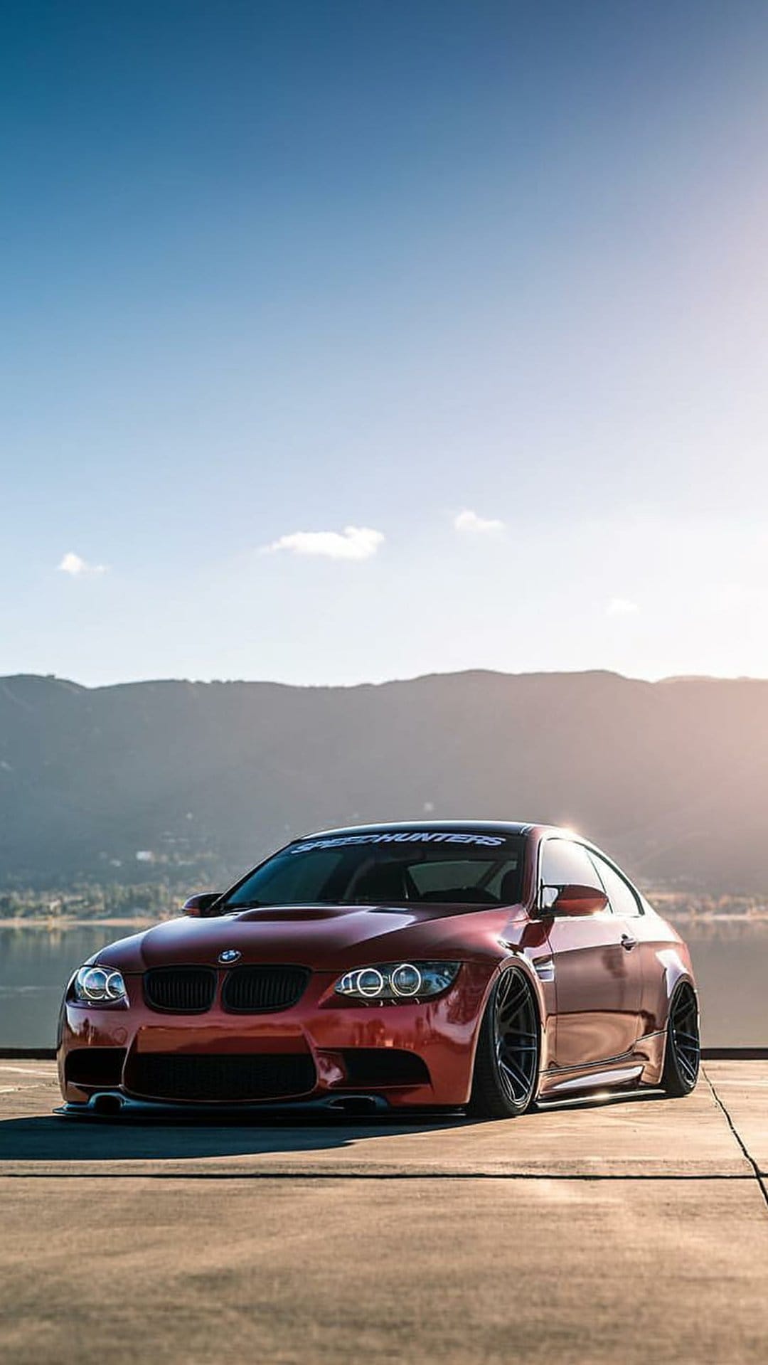 Red BMW M3 coupe with wide body fenders parked outdoors against a scenic mountain and lake horizon.