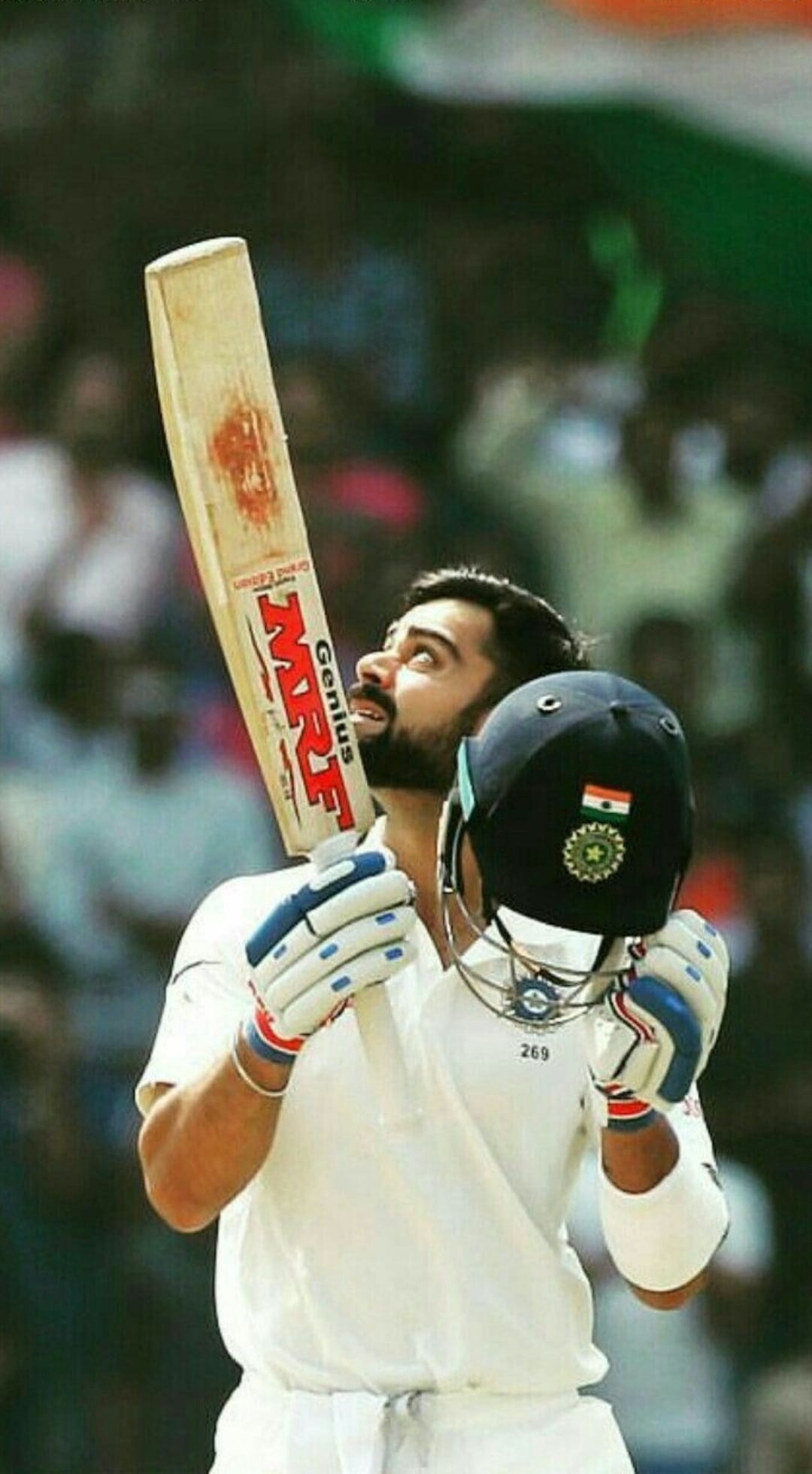 Virat Kohli raises a cricket bat and helmet with a triumphant gaze during a victory celebration on the pitch.