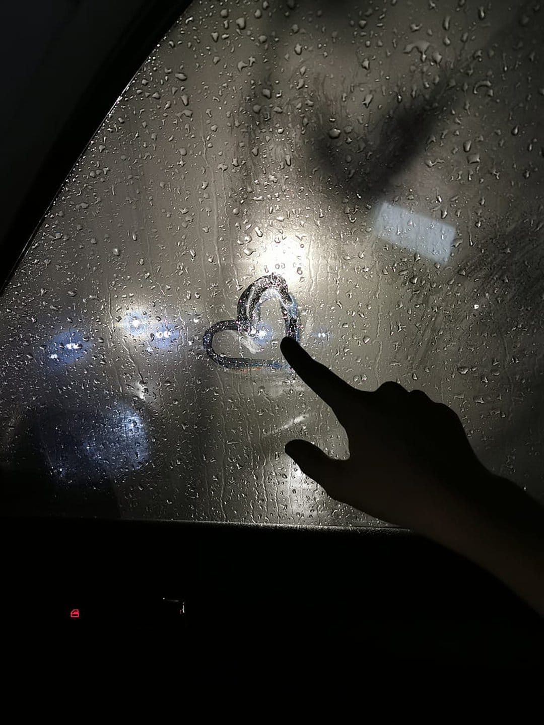 Heart shape traced on a car window covered in raindrops with blurred golden streetlights in the background.