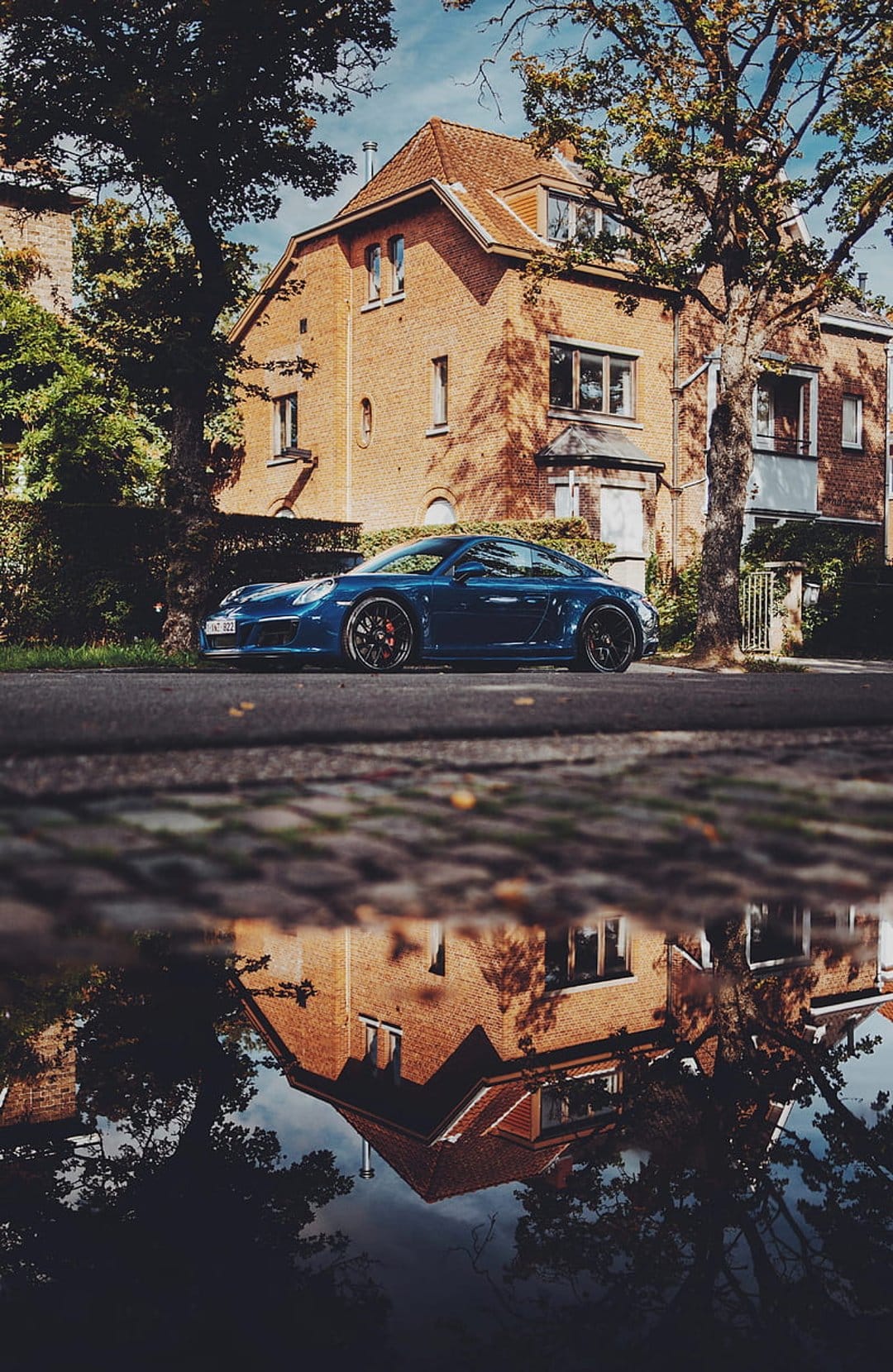 Blue Porsche sports car reflected in a rainwater puddle on a cobblestone street near a brick house and green trees.