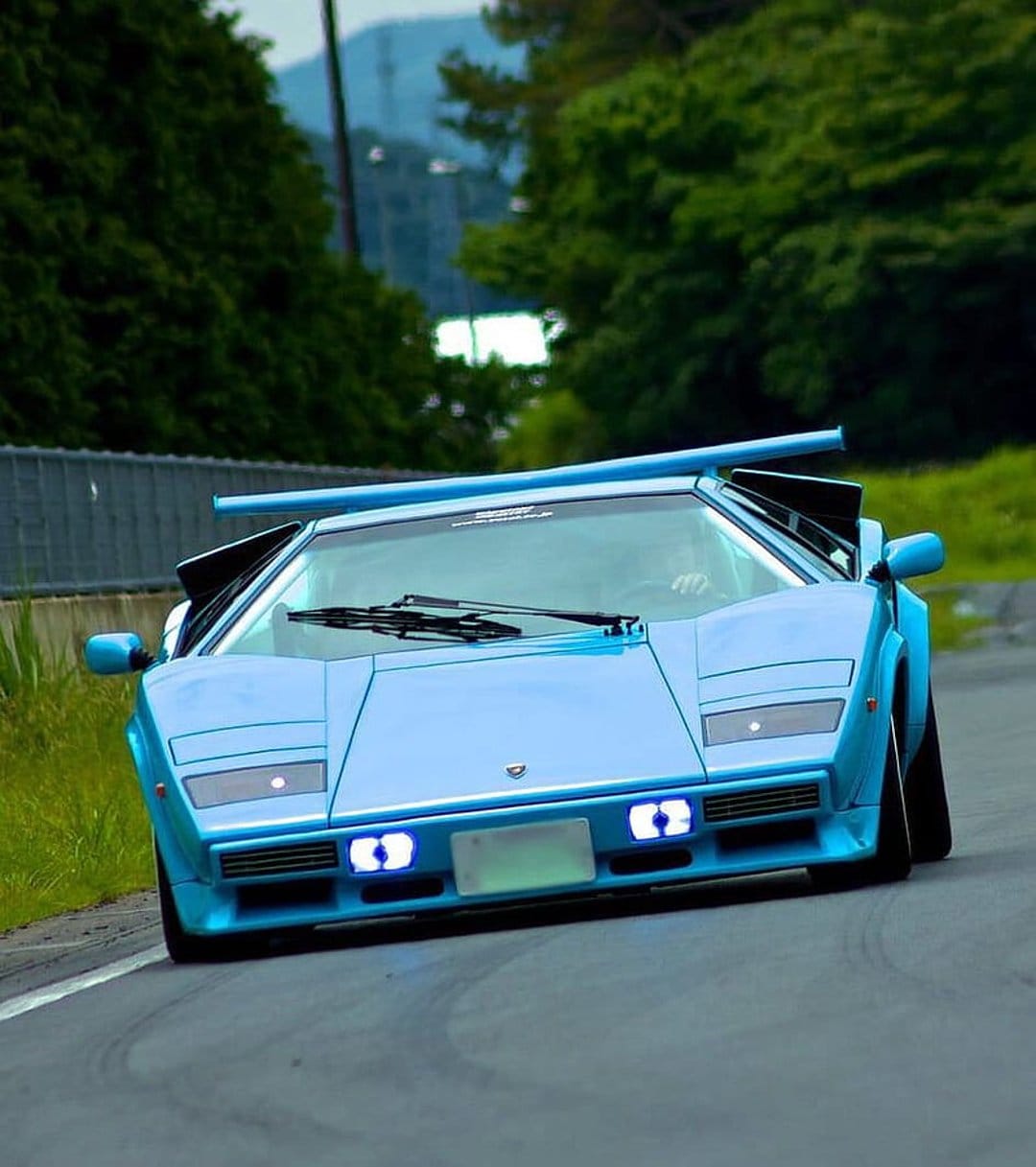 Blue Lamborghini Countach supercar leaning into a race track turn with green trees and a building behind it.