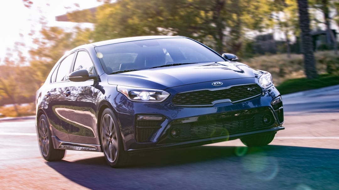 Blue Kia sedan driving on a sunlit road with motion blur on wheels and warm golden lens flares.
