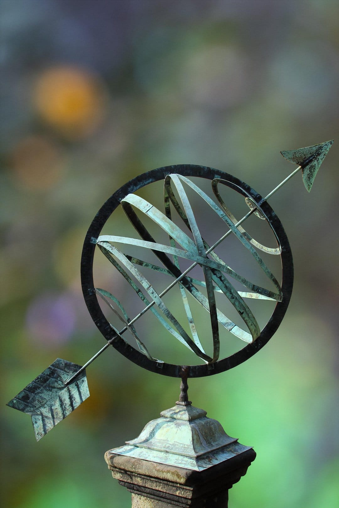Bronze armillary sphere sundial with an arrow sits on a stone pedestal against a blurred garden backdrop.