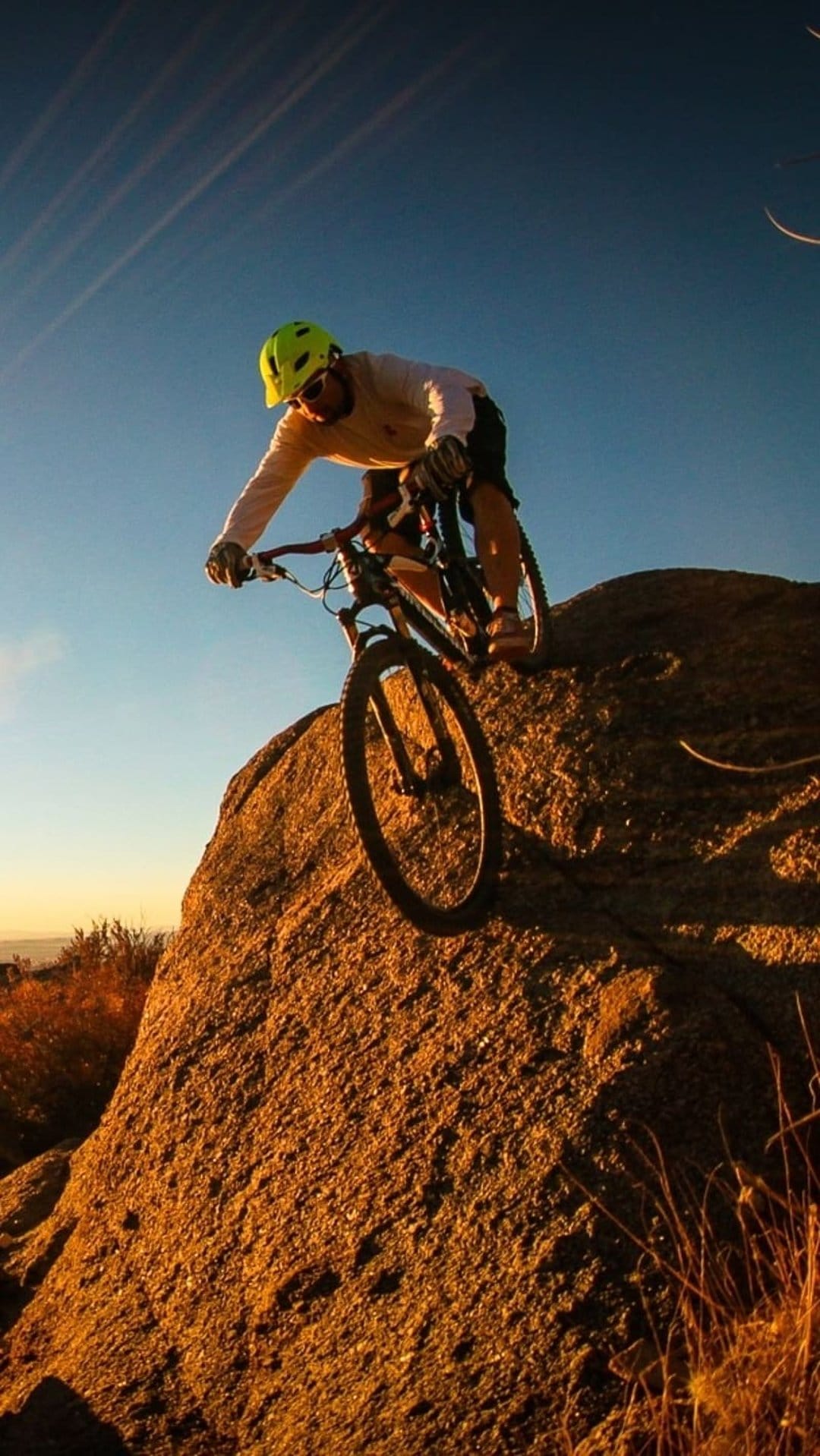 Mountain biker in a yellow helmet descends a massive rock under a warm sunset sky with deep blue horizons.