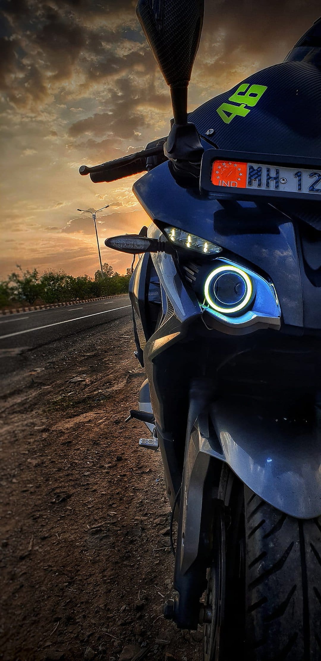 Motorcycle headlight and MH-12 license plate glow under an orange sunset sky with a background street lamp.