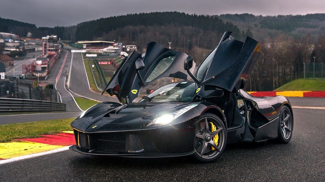 Black Ferrari LaFerrari with open butterfly doors on a wet racetrack under a moody, overcast grey sky.