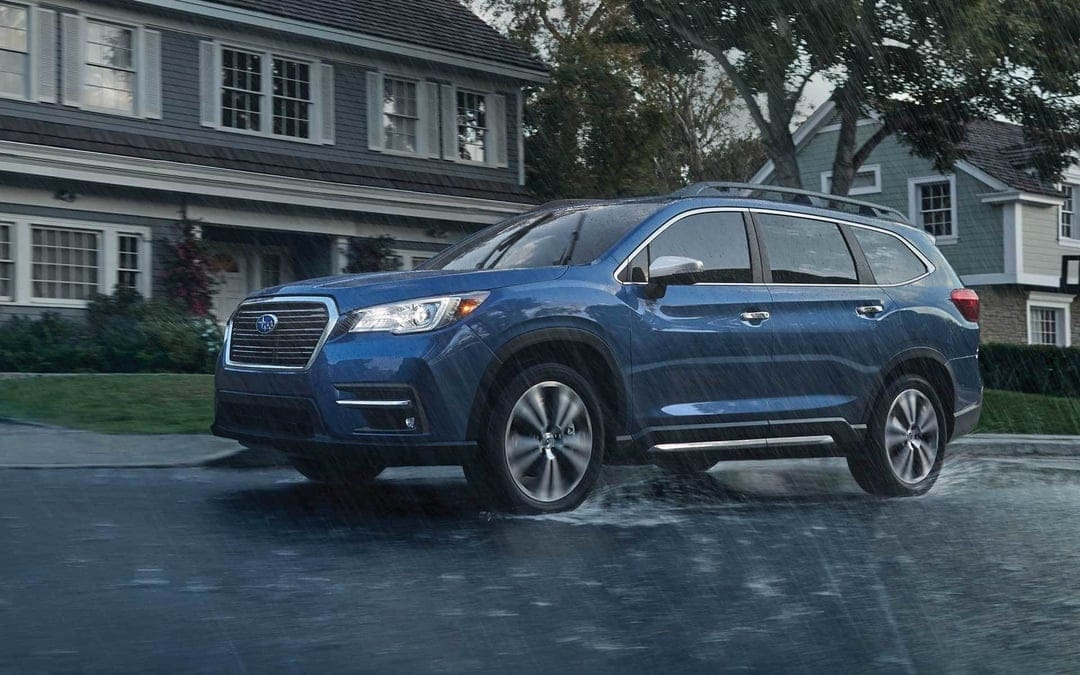 Blue SUV driving through a puddle on a rainy suburban street with houses and trees in the background.