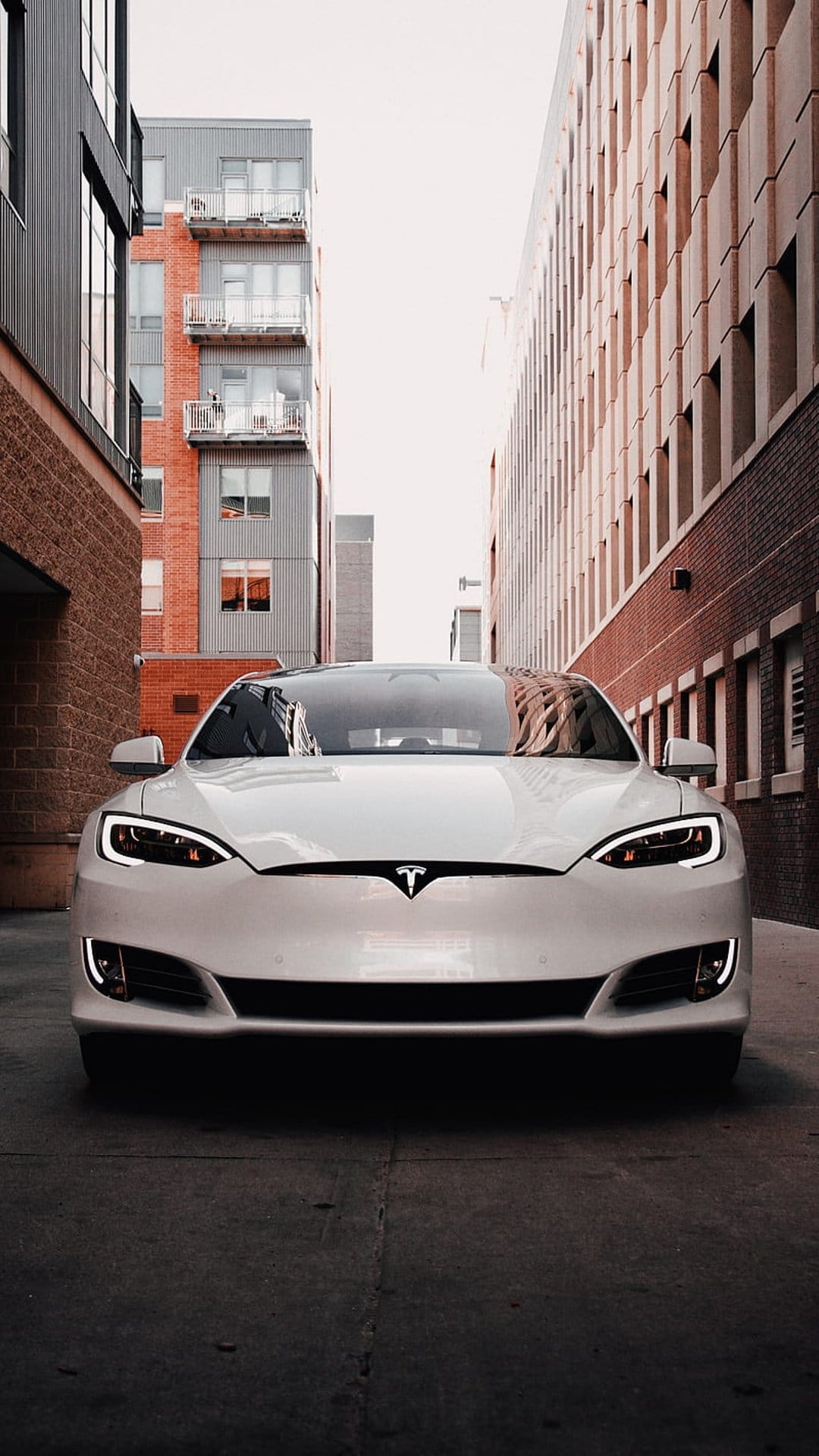 White Tesla sedan with glowing headlights parked in a narrow alleyway between industrial red brick buildings.