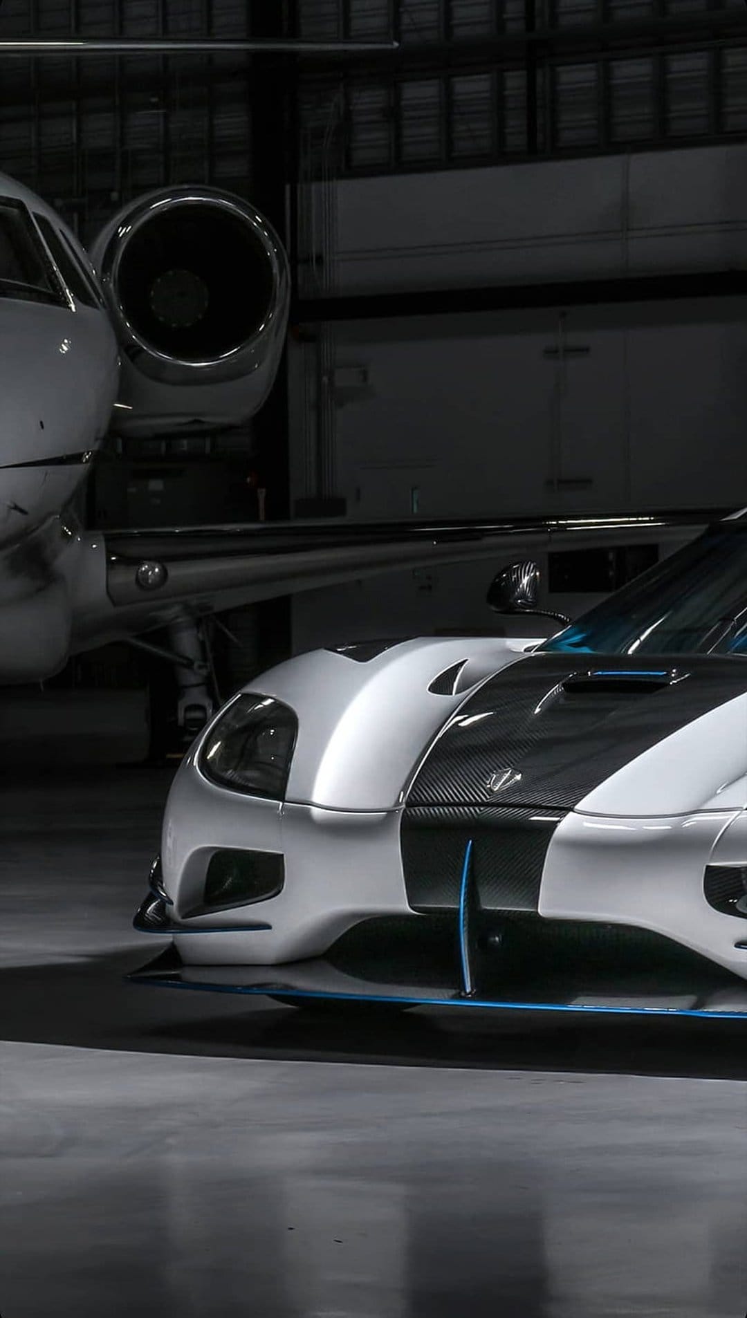 White sports car with carbon fiber accents parked next to a private jet in a dimly lit aviation hangar.