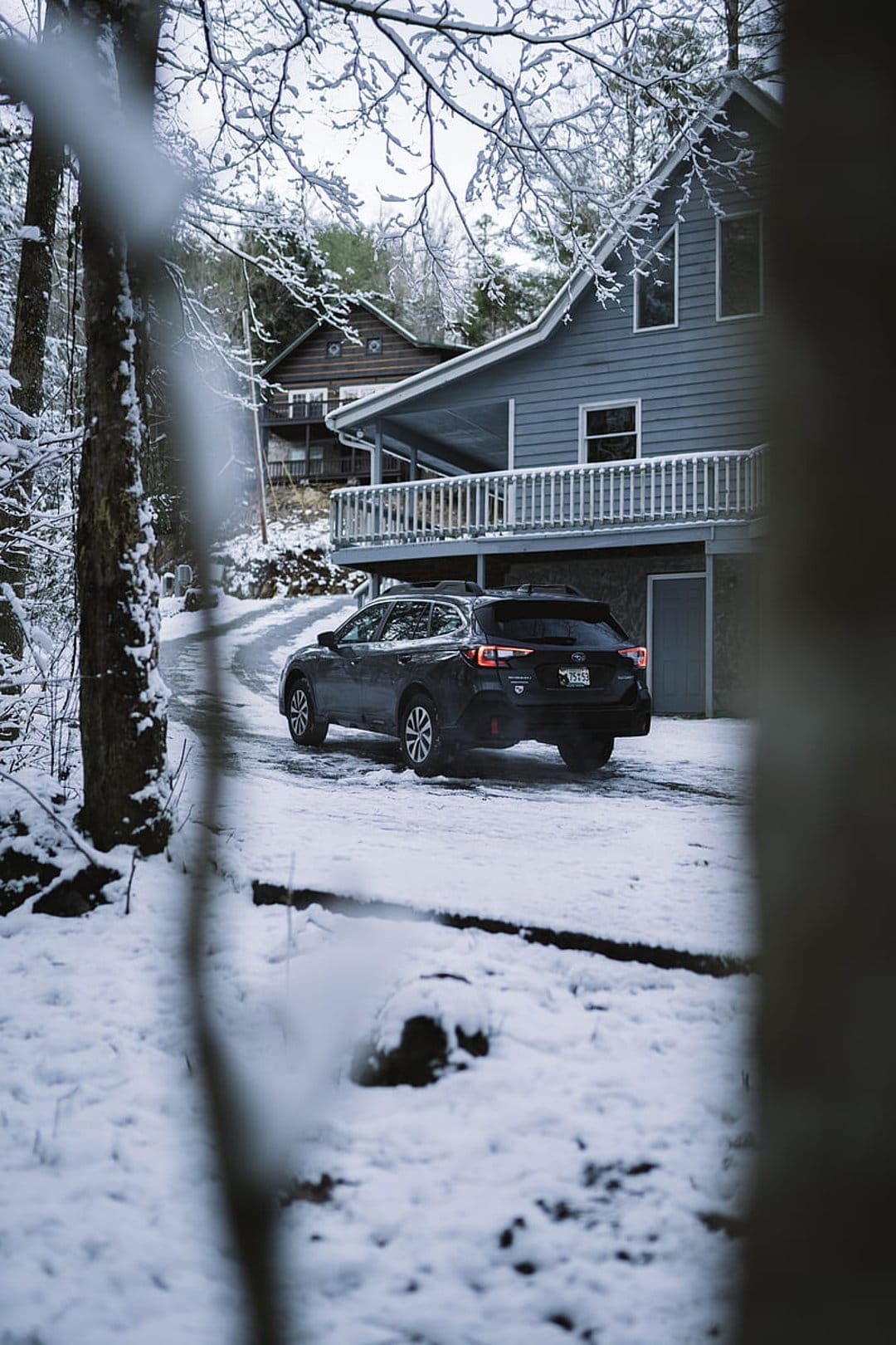 Dark SUV driving on a snowy road toward a modern gray house framed by frost-covered tree branches in a winter forest.