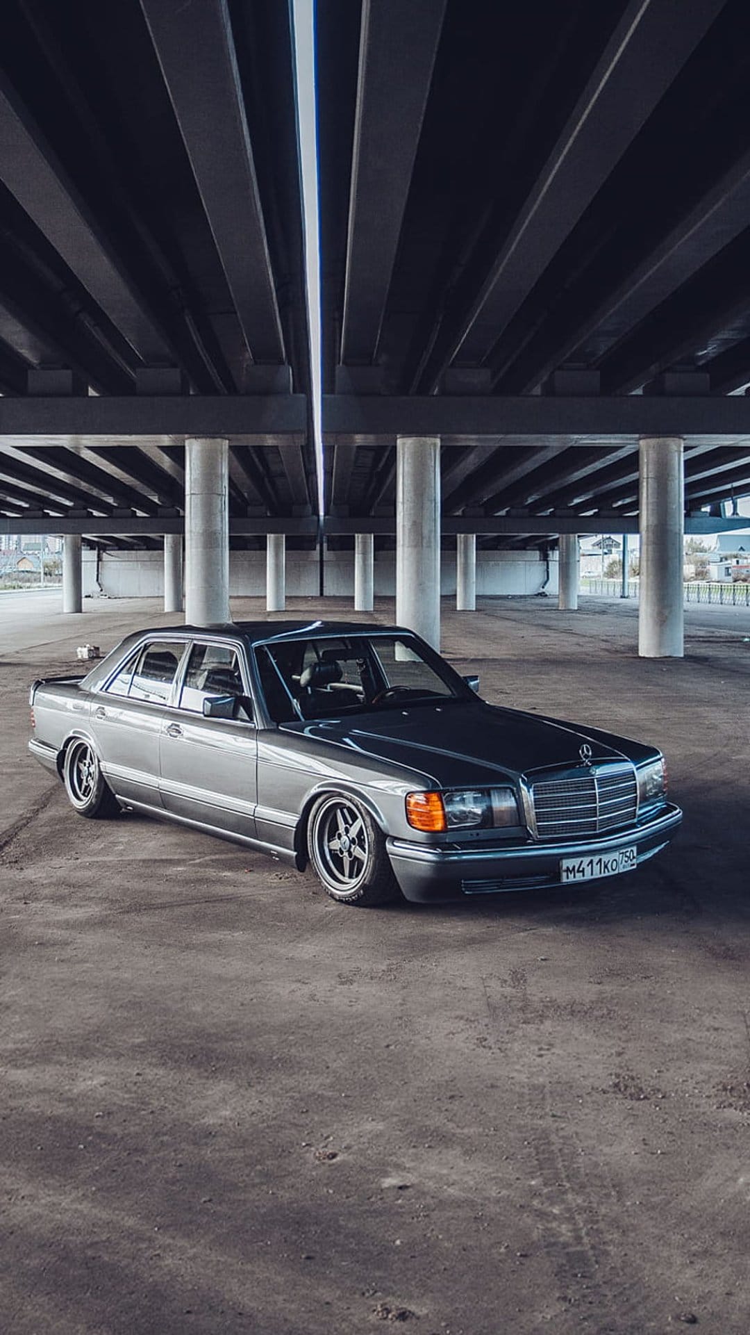 Mercedes-Benz W126 sedan with custom wheels parked on concrete under a massive grey brutalist bridge structure.