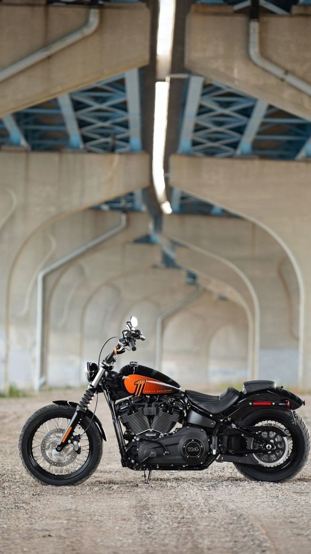 Harley-Davidson motorcycle with orange trim parked on gravel under massive concrete bridge pillars and arches.