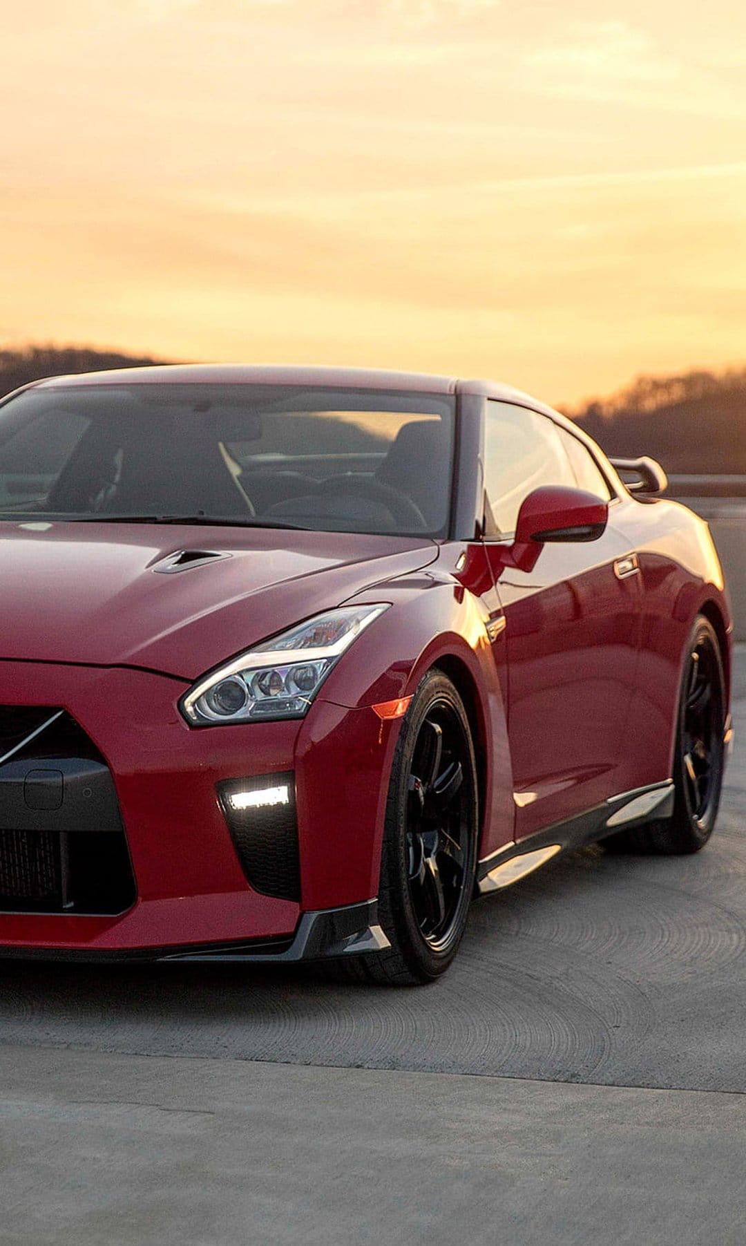 Red Nissan GT-R sports car parked on concrete with black wheels under an orange and purple sunset sky.