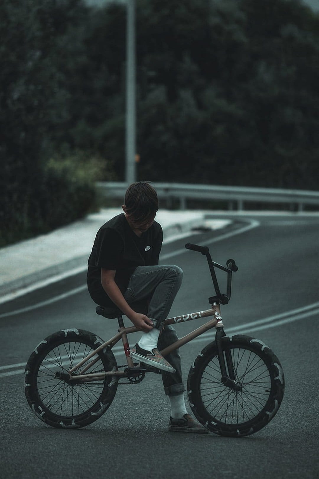 BMX rider in black clothing sits on a tan bicycle adjusting shoes on a quiet road with a guardrail behind him.