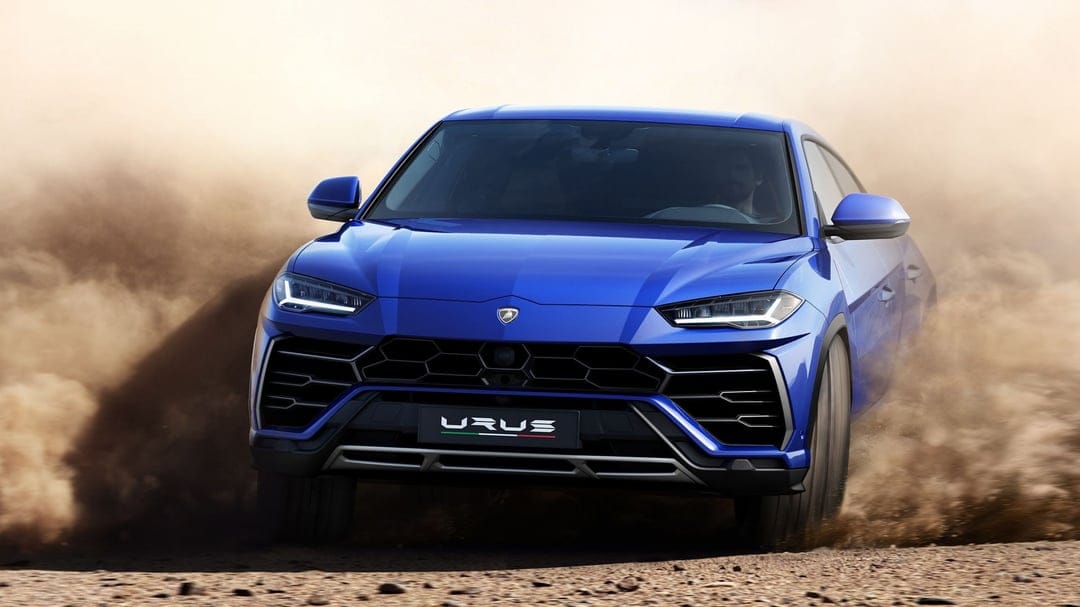 Blue Lamborghini Urus SUV driving through sand and kicking up a large cloud of dust in a desert landscape.
