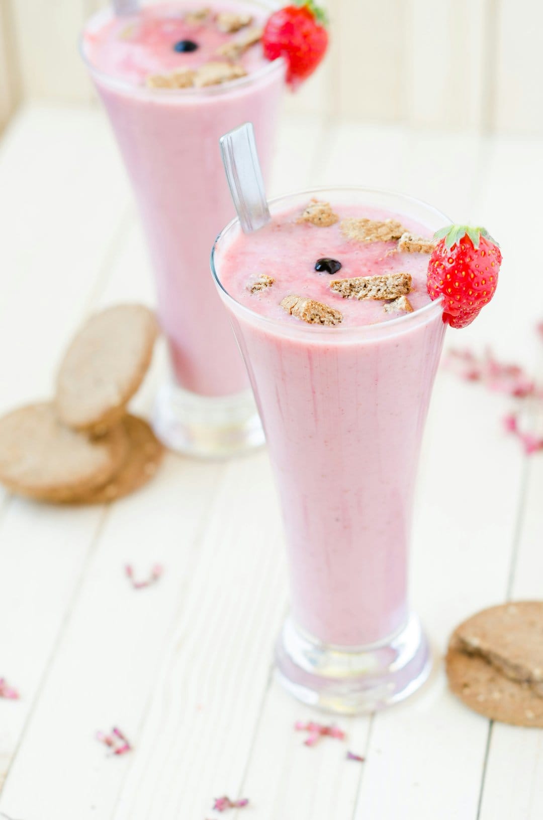 Pink strawberry smoothie in a glass with a sliced berry and cookies on a soft, light-toned surface.