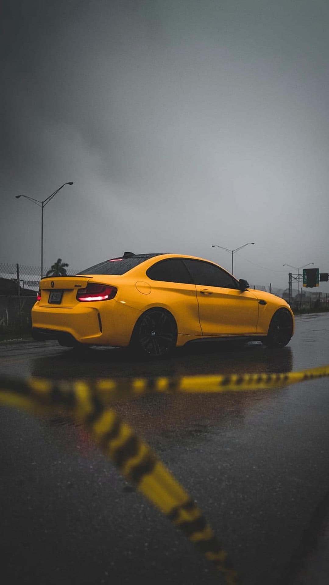 Yellow BMW M2 coupe parked on wet asphalt behind blurred caution tape under a dark, stormy sky with taillights lit.