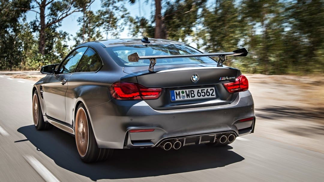 Matte grey BMW M4 GTS driving on a road with a carbon rear wing, quad exhausts, and blurred bronze wheels.