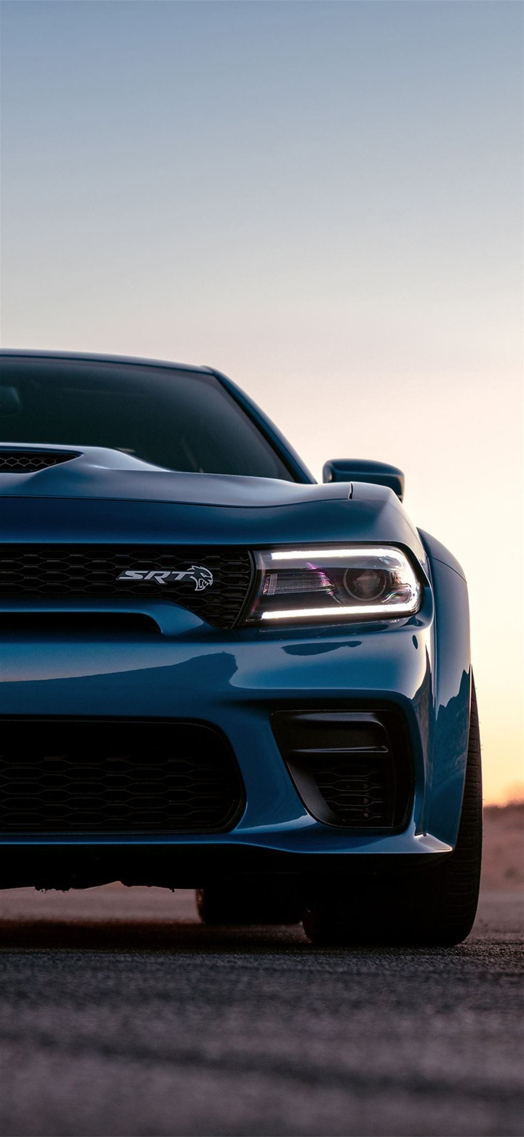 Blue Dodge Charger SRT Hellcat front end with glowing LED headlights against a soft sunset sky background.