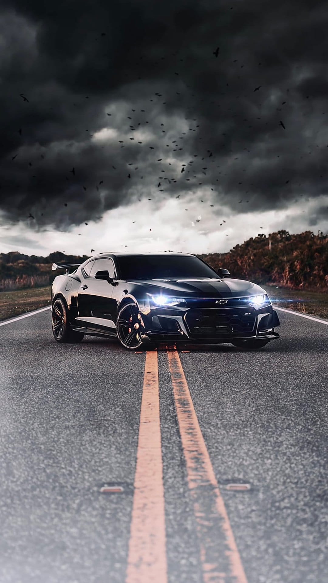 Black Chevrolet Camaro parked on a wet road with glowing headlights under a dark stormy sky with flying birds.