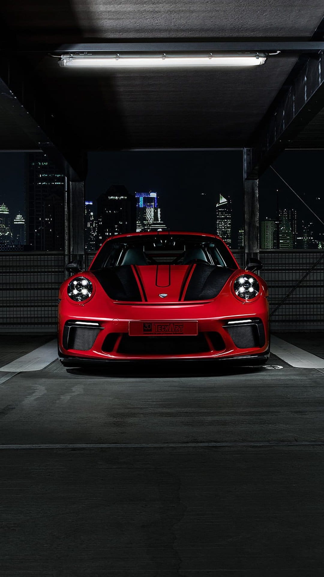 Red Porsche 911 GT3 RS with black stripes in a parking garage with a blurred night city skyline background.