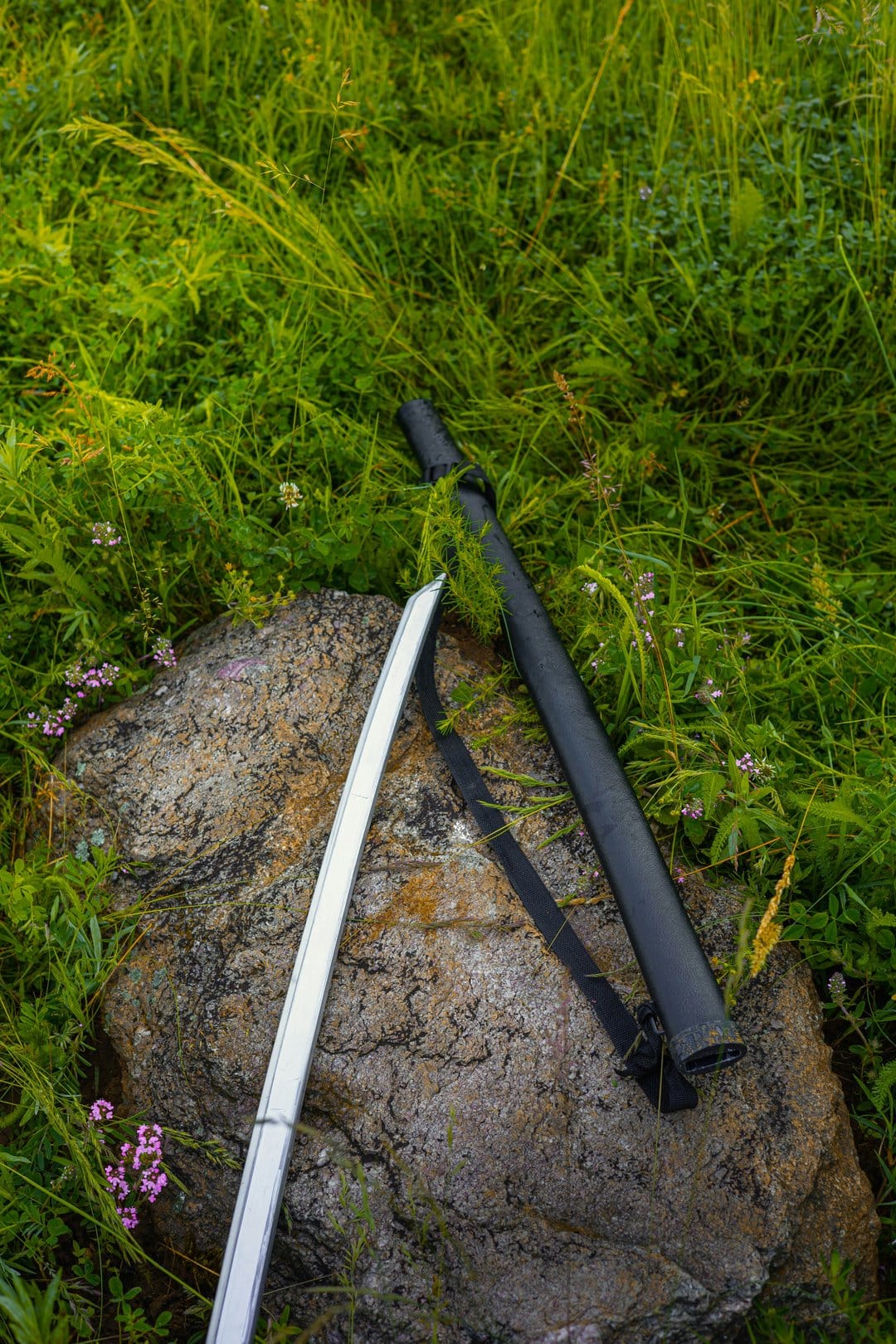 Japanese katana and white scabbard on a weathered rock surrounded by green grass and small wildflowers.