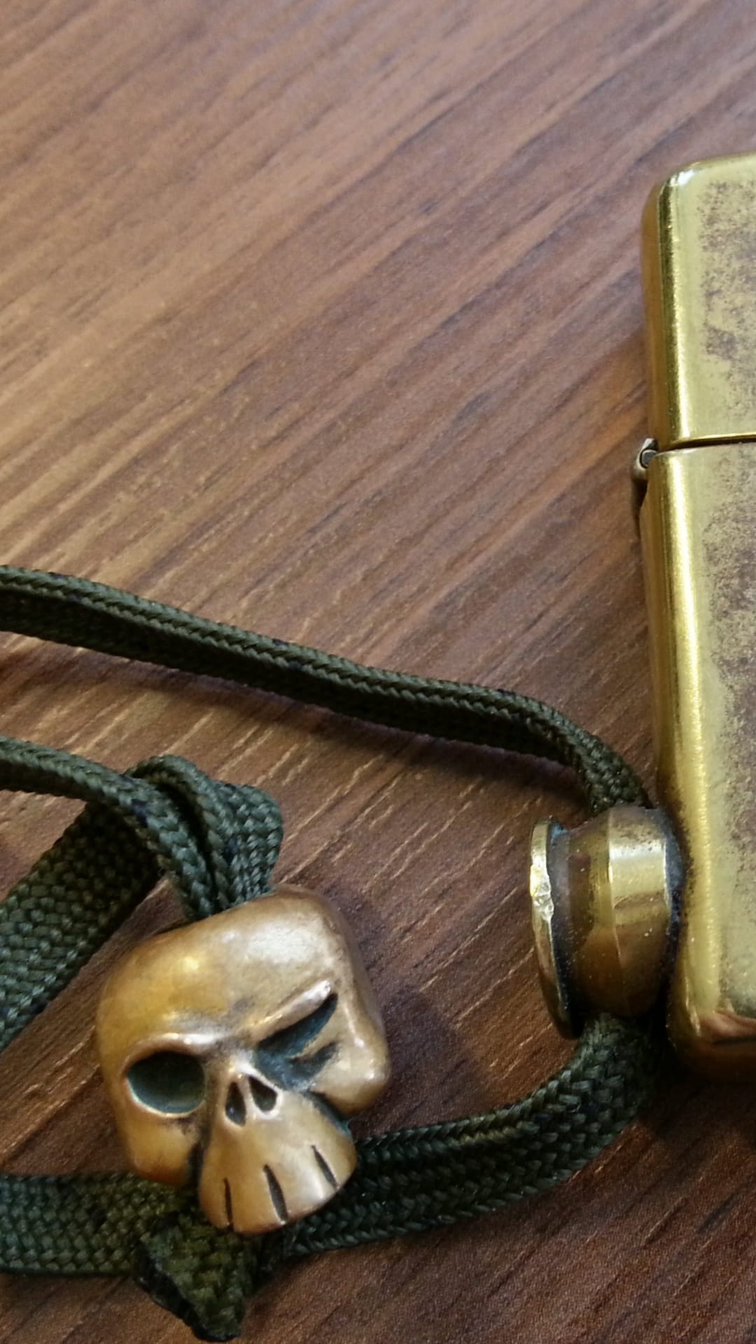 Brass skull bead and weathered metal lighter on a dark wood surface with a green braided paracord lanyard.