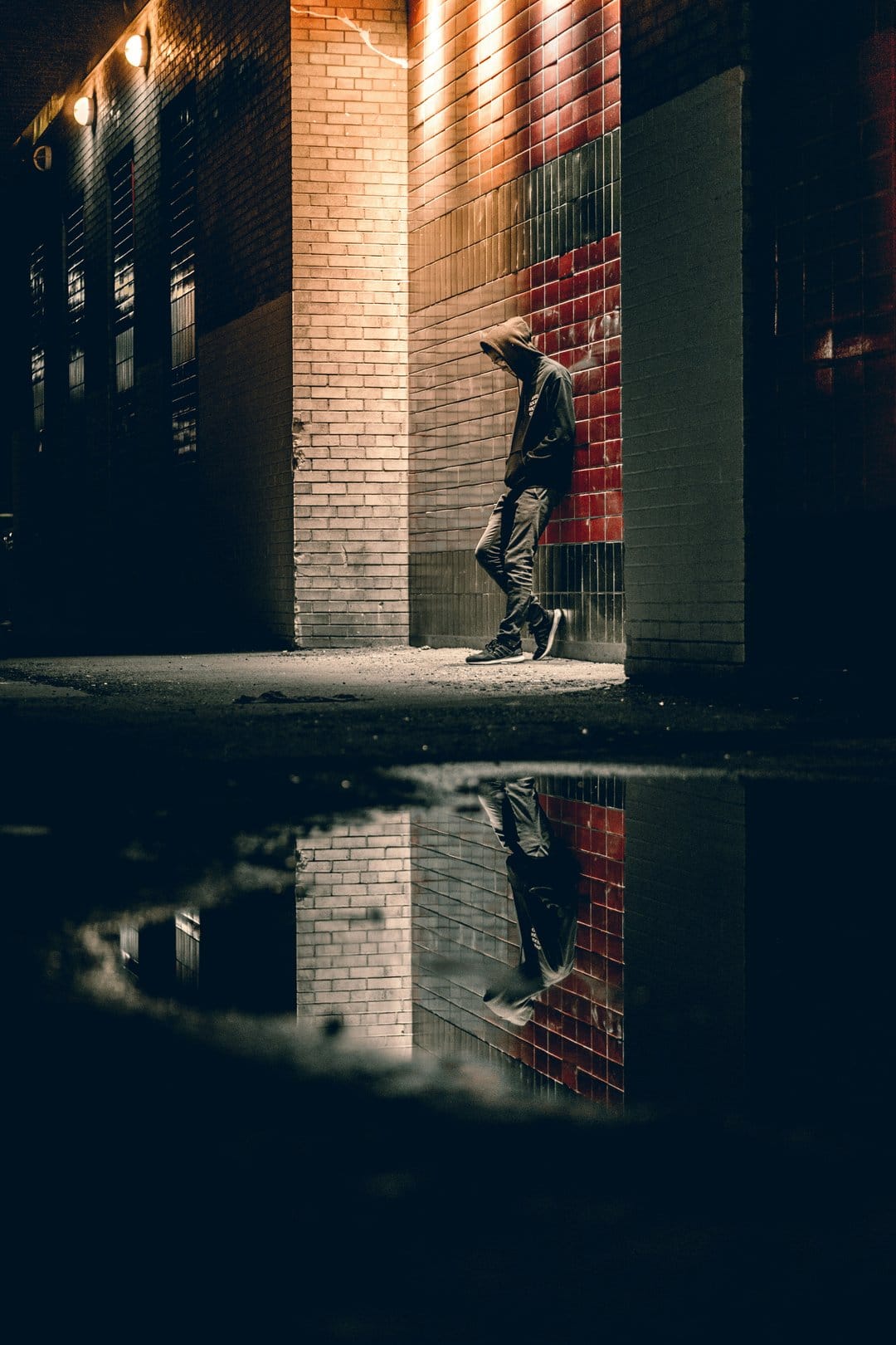 Hooded person leaning against a brick wall at night with a clear puddle reflection in a dark city alleyway.
