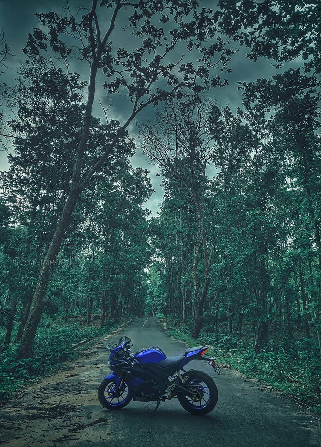 Blue sport motorcycle parked on a paved forest road beneath a dark, overcast sky with green trees lining the path.