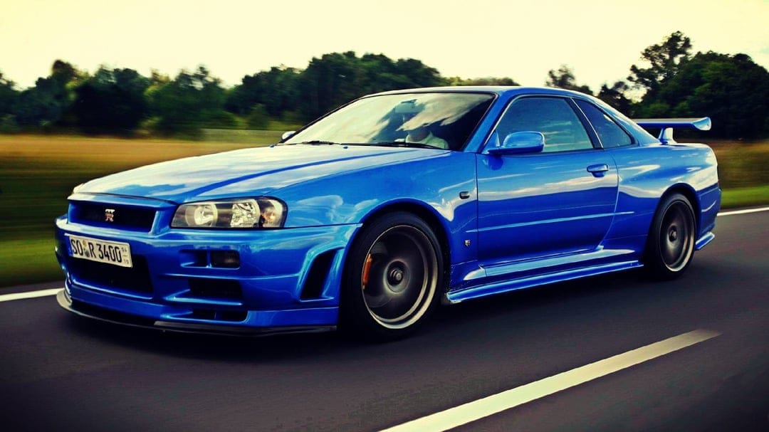 Blue Nissan Skyline GT-R R34 driving on a road with motion blurred wheels and a bright sky background.