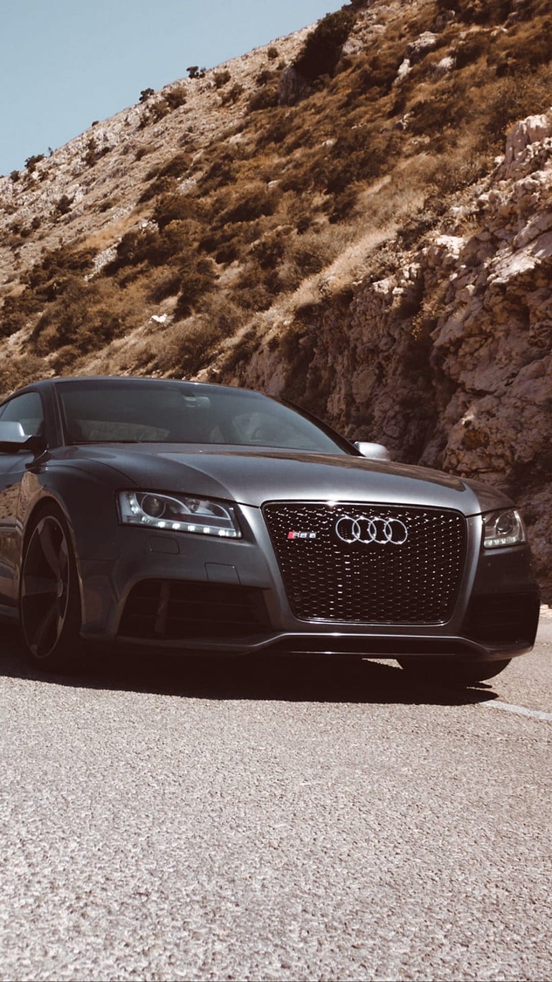 Gray Audi RS5 grille and LED headlights on a paved mountain road with rugged rocky hills in the background.